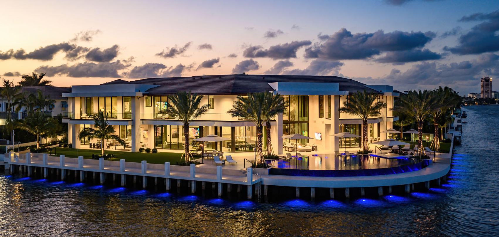 Waterfront modern estate at dusk with a lit dock and palms